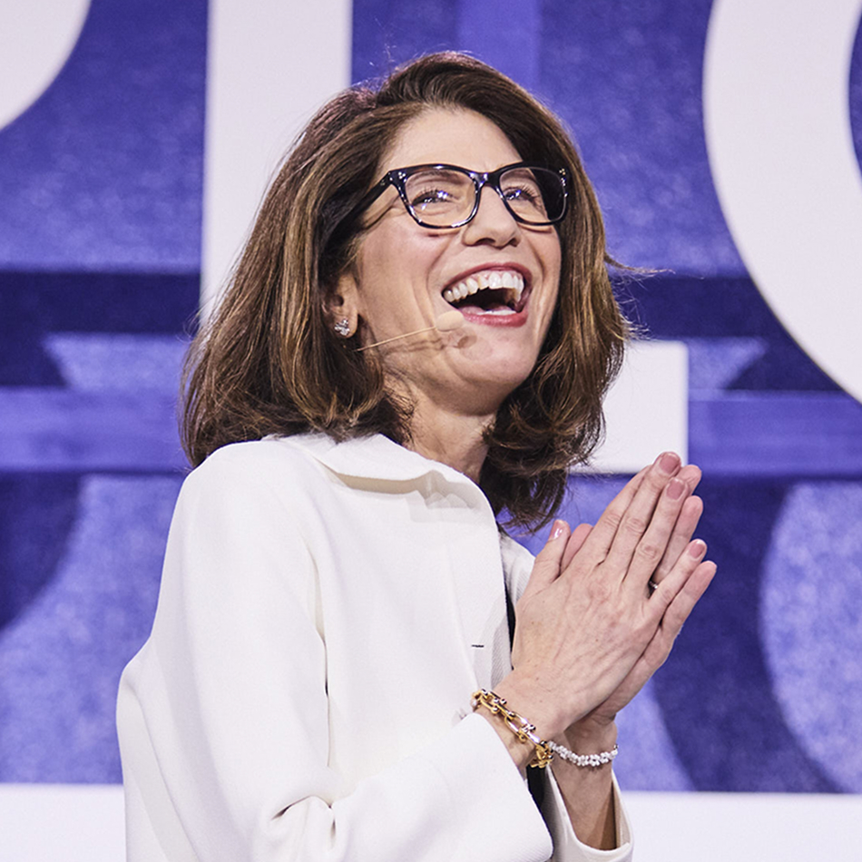 Ariane Gorin smiling on stage wearing glasses and a white jacket, speaking into a headset microphone with hands clasped in front of her, standing against a large blue abstract backdrop with oversized letter shapes.