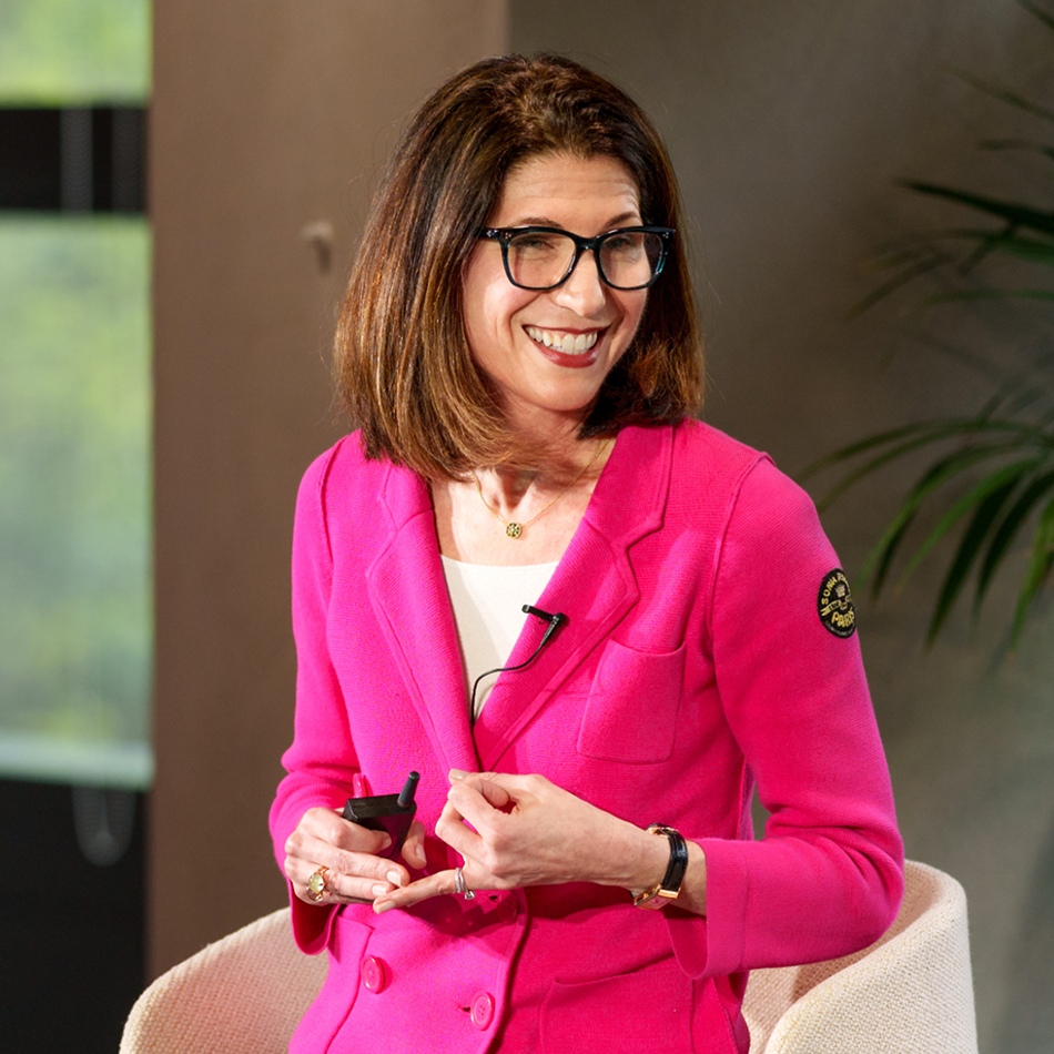 Ariane Gorin in a bright pink blazer smiles while talking on stage, holding a clicker and seated in a chair.