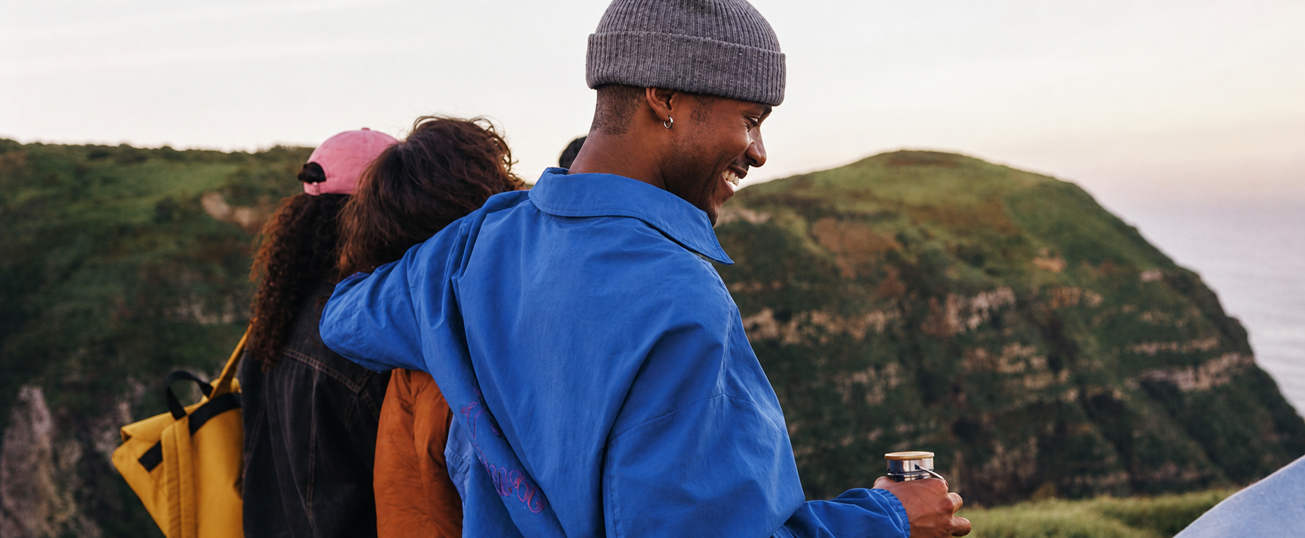 Group of friends walking along a grassy coastal trail at sunset, one person in a blue jacket with an arm around a companion while holding a travel mug, with cliffs and soft evening light in the background.