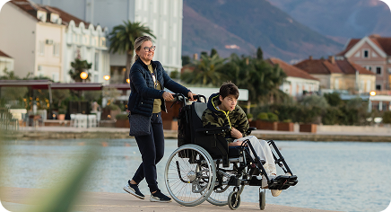 Person pushing a wheelchair user along a waterfront promenade in a coastal town.