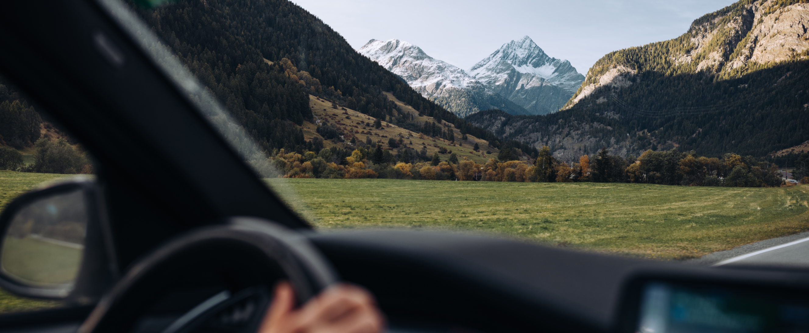 View from inside a moving car looking out through the windshield toward a wide valley of green fields, dense forested hills, and snow-capped mountains in the distance, with part of the steering wheel and driver’s hand visible in the lower foreground.