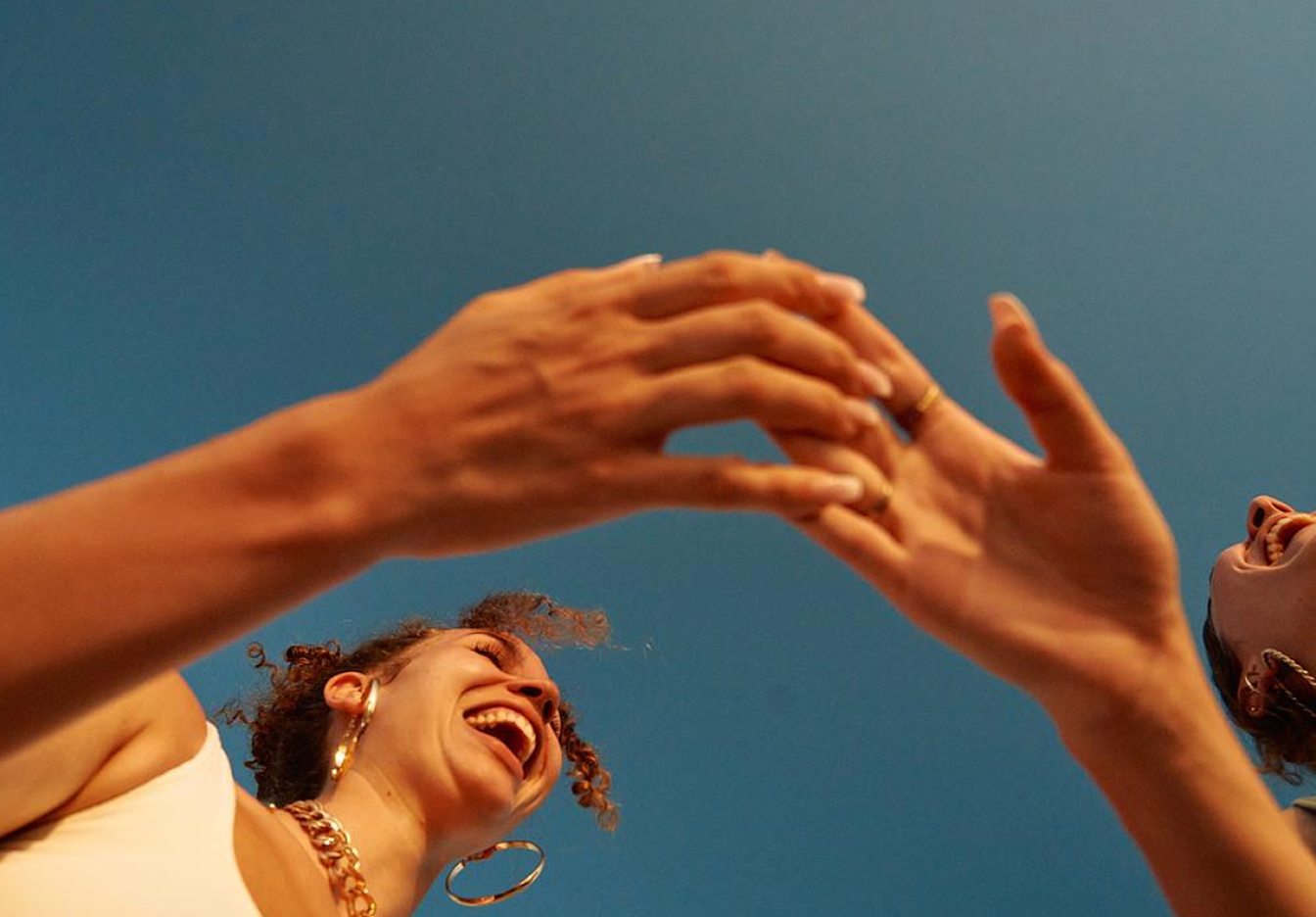 Low-angle image of two friends laughing and reaching their hands toward each other against a clear blue sky, with one person wearing large hoop earrings and a chunky necklace.