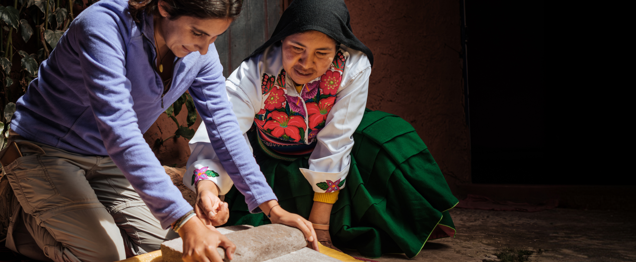 A traveler works alongside a local host to grind ingredients on a stone surface during a cultural experience.